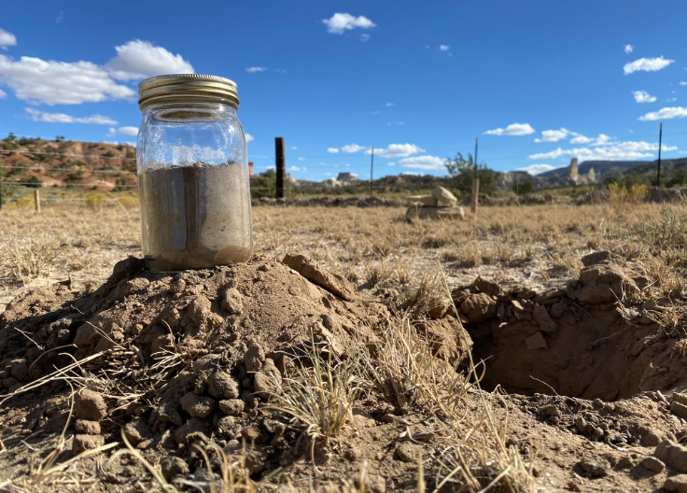 A glass jar of settled soil sitting on a mound of dug dirt beside a planting hole, fence line and mesas behind